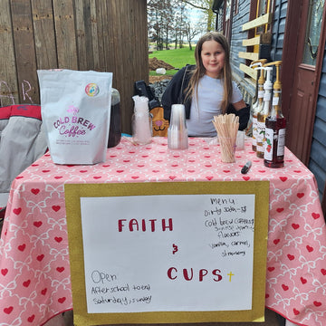 A woman stands behind a small outdoor coffee station table covered with a pink heart-patterned tablecloth. A large white bag labeled 'cOld Brew Coffee' sits on the table, along with cups, a bottle, and a handwritten sign reading 'FAITH & CUPS'.