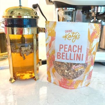 A bag of Drink Katy's Peach Bellini Kefir Tea next to a glass French press filled with brewed tea on a kitchen countertop.